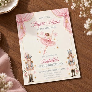 May include: A rectangular birthday invitation card featuring a ballerina in a pink tutu, two nutcracker figures, and pink curtains. The text reads: Our Little Sugar Plum Is Turning One! Isabella's First Birthday, Saturday, December 11, 3 PM.