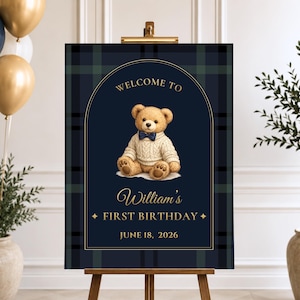 May include: A rectangular welcome sign featuring a dark navy blue and green plaid background with a gold arched border. The sign displays a teddy bear wearing a cream sweater and navy bow tie, with the text "WELCOME TO William's FIRST BIRTHDAY JUNE 18, 2026" in gold.