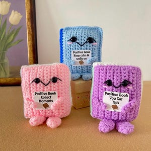 Peut inclure: Trois jouets en forme de livre crochetés en bleu, rose et violet. Chacun a des yeux en bouton noirs, un sourire cousu et de petits pieds. Le livre bleu dit "Keep calm & smile on". Le livre rose dit "Collect Moments". Le livre violet dit "You Got This".