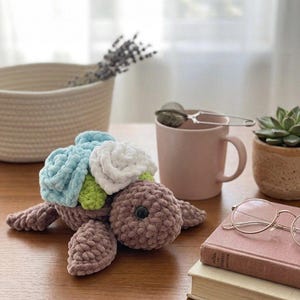 Pode incluir: Um peluche de tartaruga de crochê feito à mão, com corpo, barbatanas e cabeça marrons. A carapaça da tartaruga é adornada com flores azuis, verdes e brancas. O peluche está sobre uma superfície de madeira, com uma caneca, um livro e uma pequena planta no fundo.