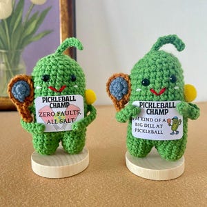 Könnte beinhalten: Zwei handgefertigte grüne Pickleball-Figuren aus Häkelarbeit. Jede Figur hält einen kleinen Schläger und einen gelben Ball. Eine Figur hat ein Schild mit der Aufschrift "ZERO FAULTS, ALL SALT". Die andere sagt "I'M KIND OF A BIG DILL AT PICKLEBALL."
