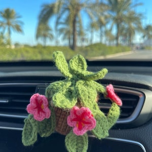 Könnte beinhalten: Eine handgefertigte Häkelpflanze mit grünen Blättern und rosa Blüten in einem braunen Topf. Die Pflanze befindet sich auf dem Armaturenbrett eines Autos. Im Hintergrund sind Palmen und ein klarer blauer Himmel zu sehen.