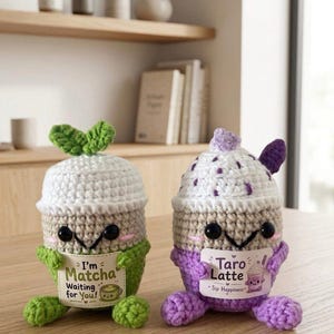 May include: Two crocheted plushies shaped like beverage cups. The left one is green and white with a green leaf topper and a sign reading I'm Matcha Waiting for You. The right one is purple and white with a purple topper and a sign reading Taro Latte Sip Happiness.