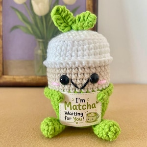 Könnte beinhalten: Ein gehäkeltes Plüschtier in Form eines Matcha-Getränks mit weißer Oberseite, hellgrünem Körper und zwei grünen Blättern oben. Es hat schwarze Knopfaugen, rosa gestickte Wangen und hält ein weißes Schild mit der Aufschrift I'm Matcha Waiting for You!