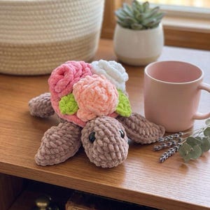 Pode incluir: Uma tartaruga de crochê feita à mão com um corpo e membros macios e texturizados. A carapaça da tartaruga é adornada com flores de crochê rosa, pêssego e branco, com folhas verdes. A tartaruga está em uma superfície de madeira.