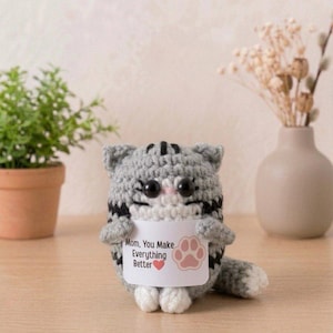 Pode incluir: Uma pequena figura de gato em crochê cinza com listras pretas, patas brancas e olhos pretos grandes. O gato segura uma placa retangular branca com os dizeres "Mom, You Make Everything Better", um coração vermelho e uma pata rosa.