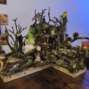 May include: A detailed fairy garden diorama with a gnome wearing a green hat with flower details, a yellow bird, and miniature mushrooms. The scene is crafted from wood, moss, and branches, creating a natural, miniature woodland scene. The base is wooden.