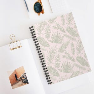 May include: A spiral-bound notebook with a pink cover featuring a repeating pattern of green botanical illustrations. The notebook is open, revealing blank white pages. Gold-colored paper clip and sunglasses are also visible.
