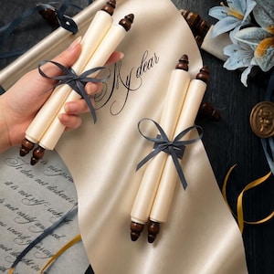 Könnte beinhalten: Cremefarbene Schriftrolle mit dunkelbraunen Holzabschlüssen, gebunden mit einem grauen Band. Die Worte "My dear" sind in eleganter Schrift auf der Schriftrolle geschrieben. Dekorative Blumen und Bänder im Hintergrund.
