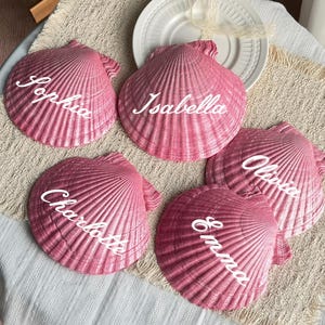 May include: Five pink scallop shell place cards with white script names: Sophia, Isabella, Charlotte, Emma, and Olivia. The shells are arranged on a textured beige surface, with a white plate and ribbon in the background.