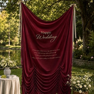 May include: A burgundy velvet wedding backdrop with the words "Happy Wedding" and a romantic quote in white script. The backdrop is draped and tied with white ribbons, set outdoors with flowers and a table.