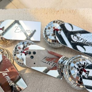 May include: A collection of personalized disco ball-shaped name tags. Each tag features a mirrored surface with a name engraved in a cursive font. The names include "Emma", "Grace", "Lily", "James", and "Emily". The tags are attached to a red string.
