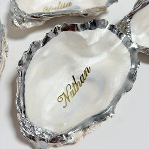May include: Several oyster shells with silver-colored edges and pearlescent interiors are displayed. The name "Nathan" is written in gold script on one shell, and "Yuliia" on another. These shells could be used as decorative dishes or place card holders.