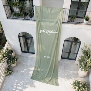 Op de afbeelding: Een lange, saliegroene stoffen banner die verticaal aan een balkonreling hangt. De banner is voorzien van witte tekst met "The Haffmann" in een cursief lettertype, gevolgd door de tekst "est 2026" in een eenvoudig, strak schreefloos lettertype.