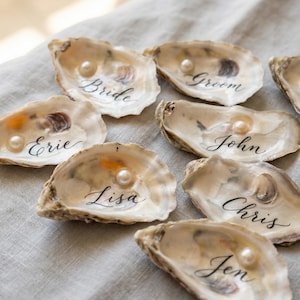 May include: Oyster shells with calligraphy names and a pearl in each shell. Names include "Bride", "Groom", "Erie", "John", "Lisa", "Chris", and "Jen". The shells are arranged on a neutral-colored surface.