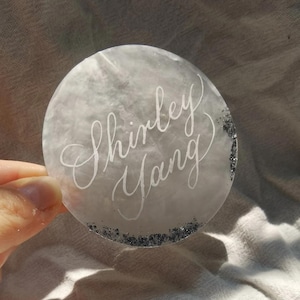 May include: A round, translucent coaster with the name "Shirley Yang" written in elegant white script. The coaster has a marbled, gray appearance with a border of silver glitter.