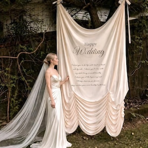 May include: A bride in a white satin wedding dress and veil stands beside a cream fabric backdrop. The backdrop displays "Happy Wedding" and a romantic quote. The scene is set outdoors with greenery and a tree.