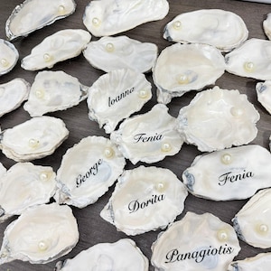 May include: A collection of white oyster shells, each with a small pearl and personalized name in black script. The shells are arranged on a dark surface, creating a unique and elegant display for place settings or event decor.