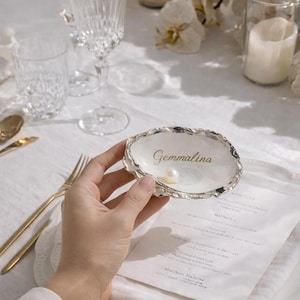 Può includere: Un piatto decorativo a forma di conchiglia con una perla e il nome "Gemmalina" in oro. Il piatto ha un bordo argentato ed è tenuto sopra una tovaglia bianca con un menu e posate dorate.