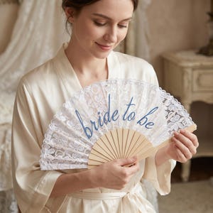 Könnte beinhalten: Ein weißer Spitzenfächer mit einem hellen Holzrahmen. Der Fächer zeigt den Schriftzug "bride to be" in blauer Schreibschrift in der Mitte des zarten Blumenmusters. Der Fächer ist halbkreisförmig und wird in geöffneter Position gehalten.