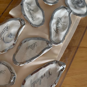 May include: Several painted oyster shells, each inscribed with a name in cursive script. The shells feature a white interior, a silver-painted edge, and are displayed on a light-toned surface. Names include "Katherine", "Chrissy", and "Kimberly".