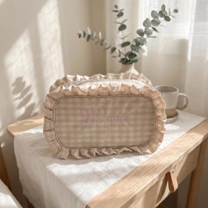 Peut inclure: Une trousse de maquillage à carreaux beiges et blancs avec un bord à volants. Le sac porte le nom "Megan" brodé en écriture cursive. Le sac est posé sur un tissu blanc sur une table de chevet en bois.