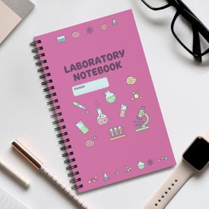 Puede incluir: Un cuaderno de laboratorio rosa con espiral con las palabras "LABORATORY NOTEBOOK" impresas en la portada. La portada también presenta ilustraciones con temática científica y un espacio para el nombre del propietario. También se ven un bolígrafo, gafas y un reloj inteligente.