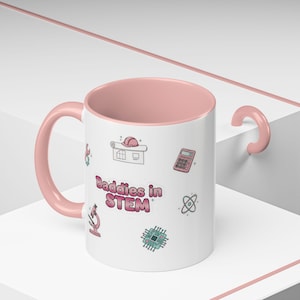 May include: White ceramic mug with a pink interior and handle. The mug features STEM-related icons and the text "Baddies in STEM" in pink. The design includes a calculator, microscope, and atom symbol.
