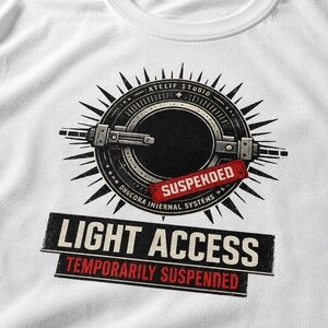 May include: A white t-shirt featuring a graphic print of a circular mechanical device with spikes. The design includes the text ATELIF STUDIO, DHECORA INIERNAL SYSTEMS, SUSPEKDED, LIGHT ACCESS, and TEMPORARILY SUSPENDED in black, white, and red ink.