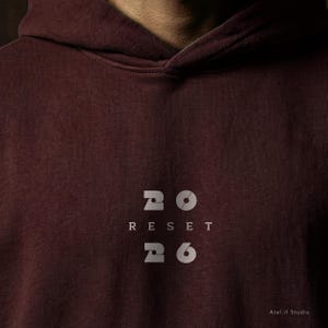 May include: A maroon hoodie with the word "RESET" and the numbers "20" and "26" printed in a light gray font. The hoodie has a drawstring hood and a ribbed texture.