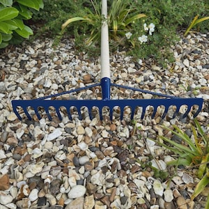 May include: A blue metal garden rake with a wooden handle. The rake's head has curved tines for gathering leaves and debris. The handle is a light beige color. The rake is resting on a bed of small stones and pebbles.