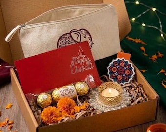 May include: A cardboard gift box containing a beige canvas pouch with an elephant print, a red card with the text Happy Diwali, three gold-wrapped Ferrero Rocher chocolates, a lit candle in a gold holder, and a colorful floral mandala sticker.