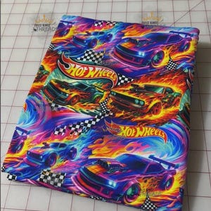 May include: Fabric with a vibrant Hot Wheels design. The fabric features stylized race cars with flames, set against a backdrop of swirling blues, purples, and oranges. The Hot Wheels logo and checkered flags are also visible.