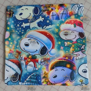 May include: A square fabric featuring Snoopy in various Christmas scenes. Snoopy is wearing Santa hats and festive outfits, surrounded by Christmas lights, stars, and gifts. The background is a mix of blue, green, and white.
