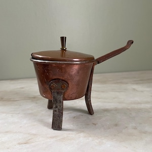 May include: A small copper pot with a matching lid and a dark brown knob handle. The pot features three sturdy metal legs and a long, angled handle extending from the side. The copper surface shows a warm, metallic finish with visible signs of age.