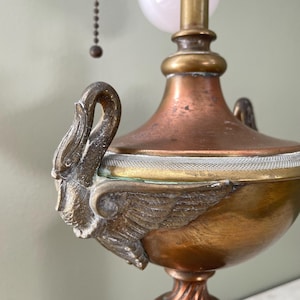 May include: An antique brass and copper lamp with swan-shaped handles. The lamp features a white globe and a pull chain. The base has a decorative, twisted design. The lamp's metal has a patina, giving it a vintage appearance.