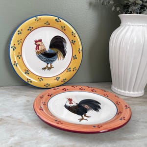 May include: Two decorative ceramic plates with rooster designs. One plate has a yellow border with blue and green accents, the other has an orange border with red and blue accents. The plates feature a white center with a colorful rooster illustration.