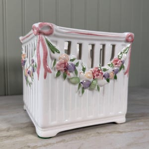 May include: A square white ceramic basket with vertical slats and a scalloped top edge. The exterior features raised, hand-painted floral garlands in shades of pink, purple, and green, accented by pink ribbons at each corner of the rim.