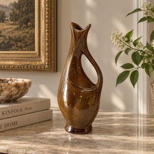May include: A tall, glossy brown ceramic vase with a sculptural, asymmetrical silhouette featuring a single integrated handle and a narrow, flared opening. The surface has a smooth, reflective finish with subtle variations in the deep brown glaze.
