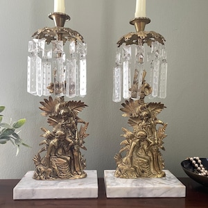 May include: A pair of brass candelabras featuring figural bases of two people, set on rectangular white marble bases. Each candelabra has a top section adorned with hanging clear glass crystals and holds a single white taper candle.