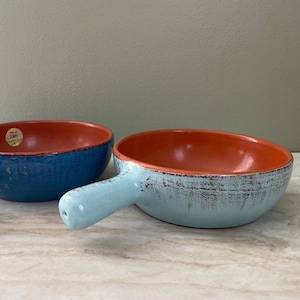 May include: Two ceramic bowls with orange interiors. One is a deep blue bowl. The other is a light blue bowl with a handle. Both bowls have a distressed, textured finish. The bowls are on a marble-like surface.