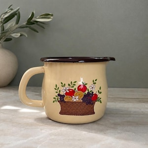 May include: A cream-colored enamel pitcher with a dark brown rim and handle. The front features a colorful graphic of a brown woven basket filled with red apples, purple grapes, and small white flowers with green leaves on a light background.