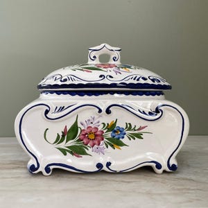 May include: A white ceramic lidded container with a curved, ornate shape and a handle on top. The piece features hand-painted blue scrollwork borders and a central floral motif in shades of pink, green, yellow, and blue on the front and lid.