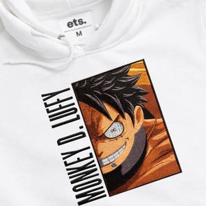 May include: White hoodie featuring a detailed embroidered image of a character's face with black hair and a toothy grin. The text "MONKEY D. LUFFY" is printed vertically on the left side. The hoodie has a hood and a tag that reads "ets. M".