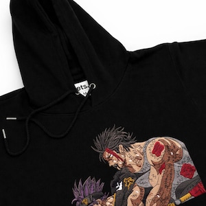 May include: Black hooded sweatshirt featuring an embroidered design of two anime-style characters in a fighting pose. The design incorporates shades of brown, red, and purple. The sweatshirt has a drawstring and a tag on the hood.