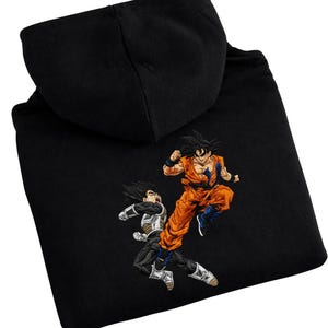 May include: Black hooded sweatshirt featuring embroidered images of two characters from the Dragon Ball Z anime series. One character is in orange and blue, the other in black and silver. The hoodie has a drawstring and a front pocket.