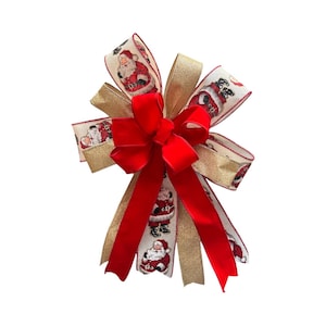 May include: A decorative bow featuring a mix of ribbons, including a cream-colored fabric with a repeating Santa Claus print, a solid red velvet ribbon, and a shimmering gold glitter ribbon, all arranged in multiple loops and long hanging tails.