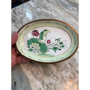 May include: An oval-shaped ceramic dish with a metallic rim, featuring a hand-painted illustration of pink lotus flowers and green lily pads on a white background, surrounded by a pale green border with dark decorative line accents.