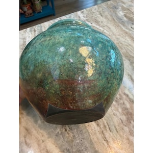 May include: A round ceramic vase with a glossy finish featuring a mottled blend of teal, green, and deep brown tones. The surface displays a textured, variegated appearance with a dark, solid-colored base and a wide, rounded body shape.