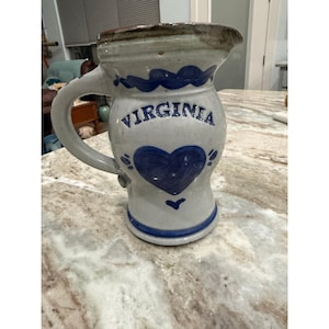 Puede incluir: Una jarra de cerámica con esmalte gris claro moteado, que presenta una línea ondulada pintada en azul en la parte superior, la palabra VIRGINIA en letras azules y un gráfico de un corazón grande en azul. La jarra tiene un asa curva y una banda azul en la base.